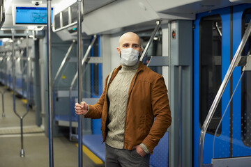 Fototapeta na wymiar A man with a beard in a medical face mask to avoid the spread of COVID-19 is riding a subway car and holding the handrail. A bald guy in a surgical mask is keeping social distance on a train.