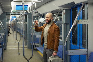 A man with a beard is taking off a medical face mask and smiling in a subway car. A bald guy with a surgical mask against COVID-19 is keeping social distance on a train.