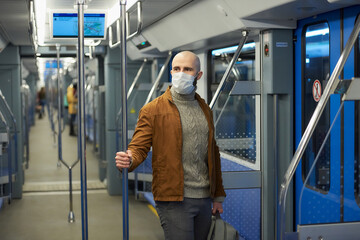 A man with a beard in a medical face mask to avoid the spread of coronavirus is riding and holding the handrail in a subway car. A bald guy in a surgical mask is keeping social distance on a train.