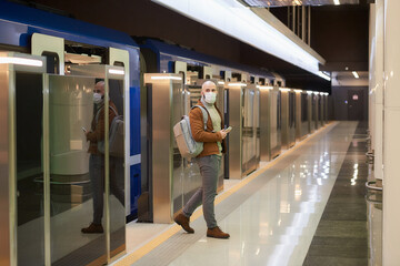 A man in a medical face mask to avoid the spread of coronavirus is holding a cellphone while leaving a modern subway car. A bald guy in a surgical mask is keeping social distance.