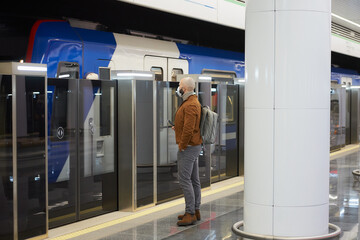 A man in a medical face mask to avoid the spread of coronavirus is holding a cellphone while waiting for a modern train on the subway. A bald guy in a surgical mask is keeping social distance.
