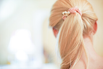 Young blonde girl with ponytail hairstyle. Pink scrunchy with pearl beads. Rear view in the room