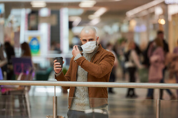 A man with a beard is putting on a medical face mask while holding a cup of coffee in the shopping center. A bald guy is keeping social distance.