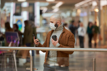 A man with a beard in a face mask to avoid the spread of coronavirus is using a smartphone and holding a cup of coffee in the shopping center. A bald guy in a surgical mask is keeping social distance.