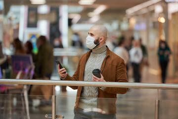 A man with a beard in a face mask is looking around and holding a smartphone and a cup of coffee in the shopping center. A bald guy in a surgical mask is keeping social distance.