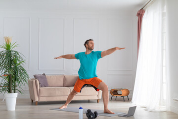 Middle aged man works out at home with an online tutorial. He performs a balance exercise in front of a laptop monitor.