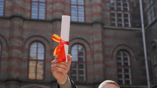 Male hand raised up holding diploma of academy graduate on the background of beautiful university school building Education concept Young graduated guy holding graduation degree convocation ceremony