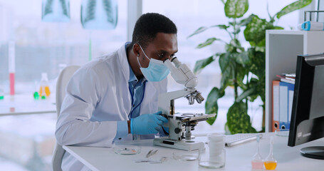 Young african medical worker in face mask examining vaccine liquid with microscope wearing gloves. Development of vaccination.