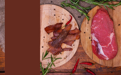 Jerky meat and raw beef on circle wooden boards on brown background with copy space. Ingredients for cooking meat snacks. Rosemary, red pepper and peppercorns. Dried meat. Top view