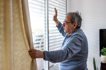 Photo of a senior man is waving from a window in his home. Happy elderly man standing by the window. Old man with eyeglasses standing by the window and looking out with raised hand, waving to someone.