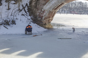 An adult man in a warm uniform is sitting and fishing on the ice near the old stone bridge