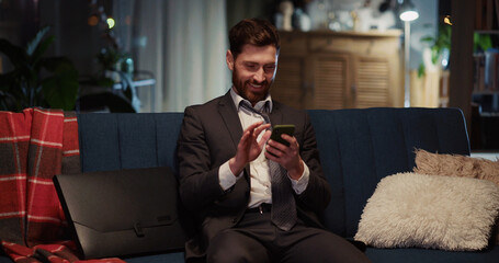 Young cheerful bearded businessman sitting at home, browsing social media on smartphone, typing text messages smiling happy. Successful people.