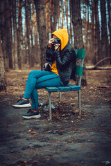 Young woman in casual clothes sitting on bench and photographing on old photo camera in forest. Female resting and taking photos with old camera in early spring.