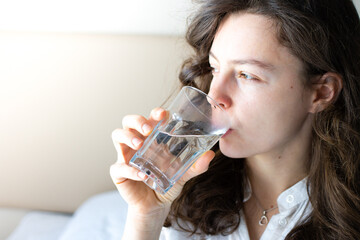 Portrait of young brunette woman drinking pure mineral water, holding transparent glass and looking in distance. Feeling thirsty after waking up. Morning routine, good healthy lifestyle habits.