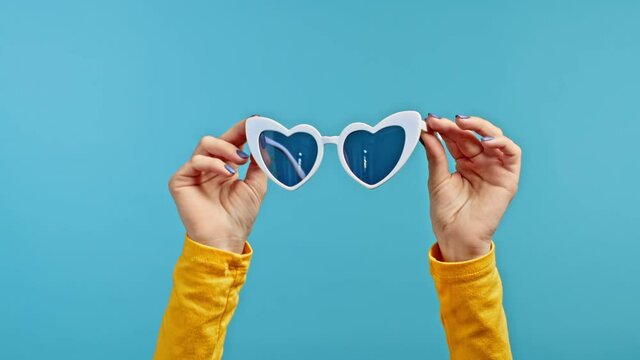 Woman's hands holding heart-shaped white sunglasses on blue studio background.