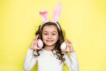Obraz na płótnie Canvas Happy curly little girl wears bunny ears, picks up colored Easter eggs, isolated on yellow background. Easter concept