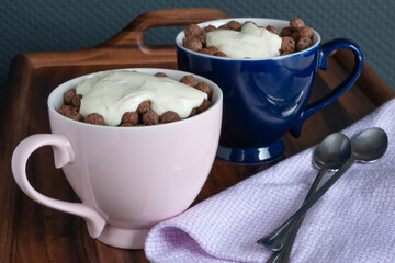 Flakes chocolate balls with yogurt in large pink and blue porcelain mugs and long little spoons wrapped in a nice cloth napkin on a wooden breakfast tray. Dry sweet breakfasts.