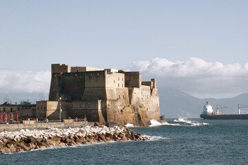 Naples Castel dell'Ovo during a storm with the waves crashing on the rocks and a cargo ship moored in the distance against the background of white clouds and Vesuvius