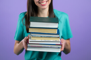 Female student holds stack of university books from library on violet background in studio. Unrecognizable woman.