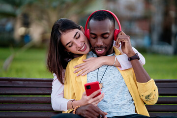 beautiful multiethnic couple in love listening to music with red headphones and smiling sitting on a city park bench, multicolored.