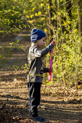 A pretty little boy on a walk in the woods, wearing a blue cap and a gray sweater, blowing soap bubbles. In the background, green trees and a well-worn path.