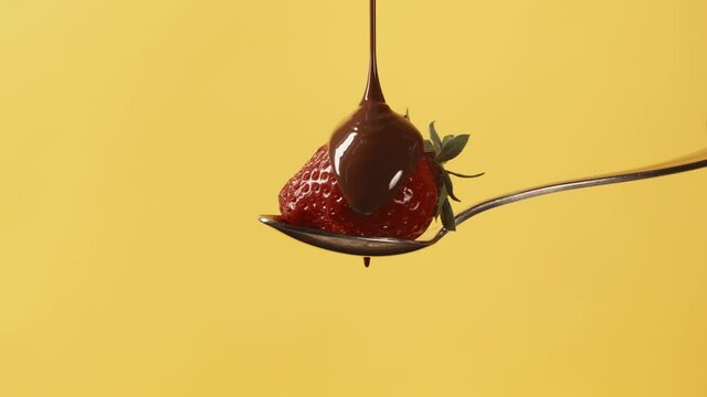 Fresh strawberry on metal pastry spoon covering with melted liquid chocolate on yellow background. Molten milk chocolate pouring on juicy red strawberry. Confectionery dessert