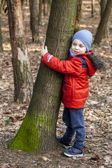 A cute little boy wearing an orange jacket, blue pants and a cap, cuddling up to a tree covered with moss. In the background, other trees and dry leaves lying on the ground.