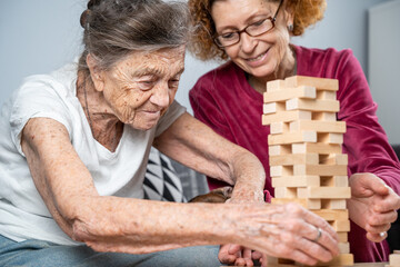Elder woman and her adult daughter together with dachshund dog spend time together at home playing board game collecting wooden blocks in tower. Jenga game. Theme is dementia and alzheimer's