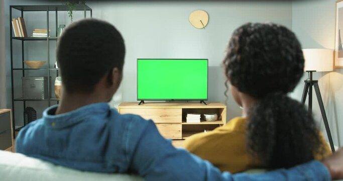 Young African American Couple Sitting At Home On Sofa And Watching Movie On Tv Talking. View From Back. Chromakey.