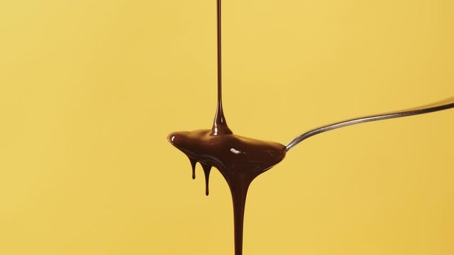 Close-up melted liquid chocolate flows, metal pastry spoon covered with flowing chocolate or brown caramel sauce stream on yellow background. Cooking sweet chocolate dessert and candies. Confectionery