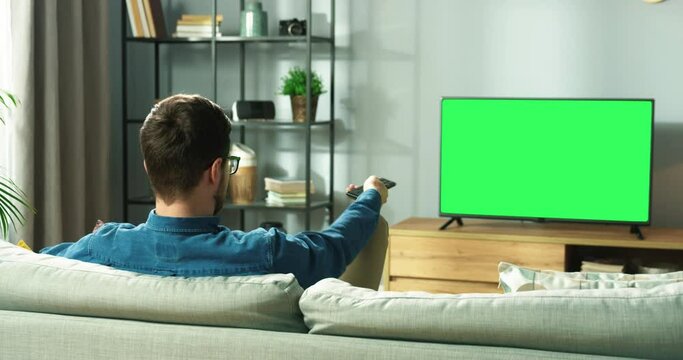 Young man sits down to relax at home on couch and using remote to find show on TV. View from back. Chromakey.