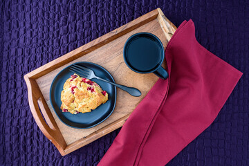 Breakfast in bed, homemade cranberry scone on black plate and wood tray on a purple bedspread

