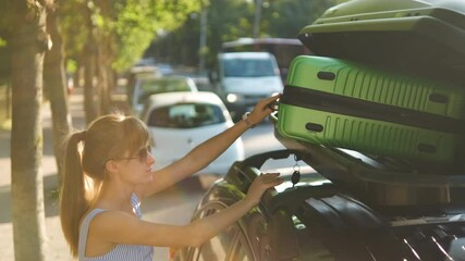 Young woman driver putting green suitcase inside car roof rack on city street. Travel and vacations concept