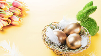 Basket easter decoration: Golden eggs in basket with spring tulips, white feathers on pastel yellow background. Congratulatory easter design.