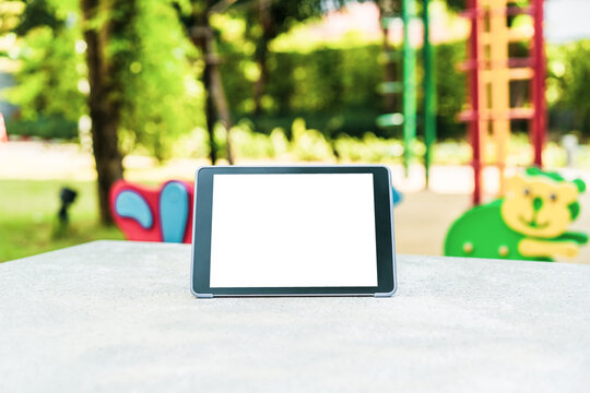 Mock-up tablet with blank white screen stands on a grey concrete table with playground background