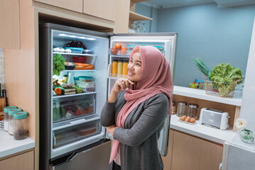 Fototapeta na wymiar muslim asian woman thinking while open the refrigerator door before cooking in the kitchen