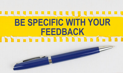 On the table are two sheets from a notebook and a pen on a yellow background written - BE SPECIFIC WITH YOUR FEEDBACK