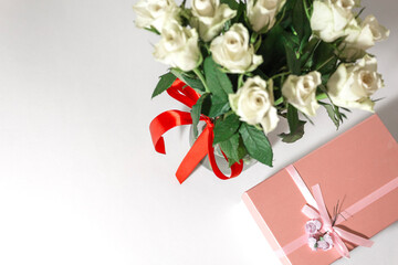 White tea roses stand in a glass vase, which is tied with a red ribbon and stands on the table. There is a gift nearby. in a pink box. Gift bouquet