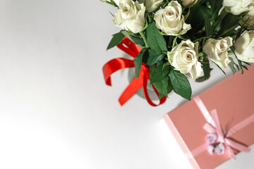 White tea roses stand in a glass vase, which is tied with a red ribbon and stands on the table. There is a gift nearby. in a pink box. Gift bouquet