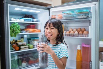 Fototapeta na wymiar thirsty happy young asian girl open fridge door drinking a bottle of water
