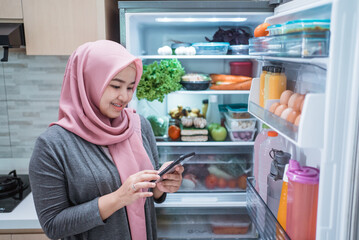 asian muslim young woman using her smartphone to buy groceries while open her fridge at home