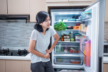 portrait of starving young asian woman hold her belly while open the fridge door looking for something to eat