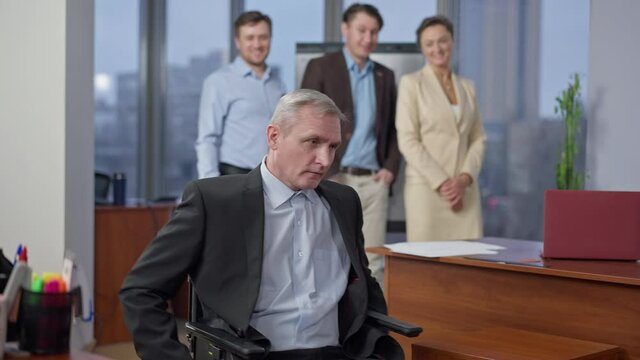 Rack focus from sad disabled stressed senior man riding away on wheelchair to group of young employees mocking at background. Frustrated paraplegic Caucasian worker leaving office