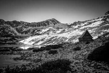 Sunny day in Tatra Mountains in black and white. Rocks covered with fresh snow and an old wooden chalet used for sheep herding. Selective focus on the building, blurred background. - obrazy, fototapety, plakaty
