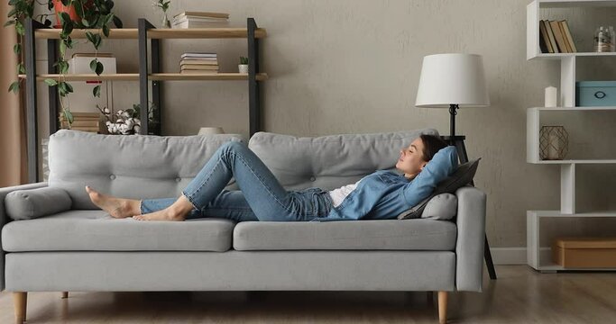 Side full length view woman with eyes closed put hands behind head lying on comfy couch breathing fresh conditioned air take break enjoy stress-free day alone in modern living room. Relaxation concept
