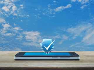 Security shield with check mark flat icon on modern smart mobile phone screen on wooden table over blue sky with white clouds, Technology internet cyber security and anti virus online concept