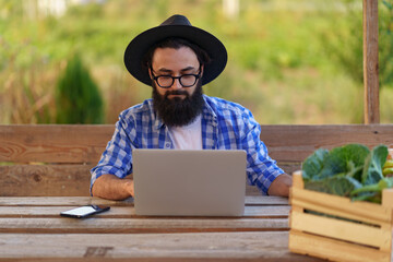 Young farmer entrepreneur sell his organic eco-friendly veggies online using laptop while sitting at a wooden table in his garden
