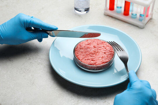 Scientist holding fork and knife over plate with minced cultured meat at light grey lab table, closeup