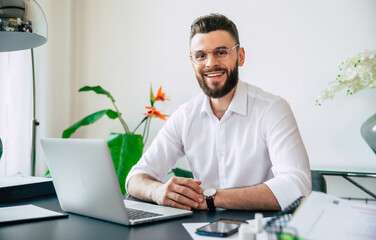 Young modern handsome stylish bearded businessman in glasses and a white shirt is working on the...