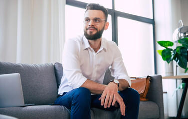 Obraz premium Confident stylish handsome bearded business man in glasses and formal smart wear is sitting on the couch in the modern office or room